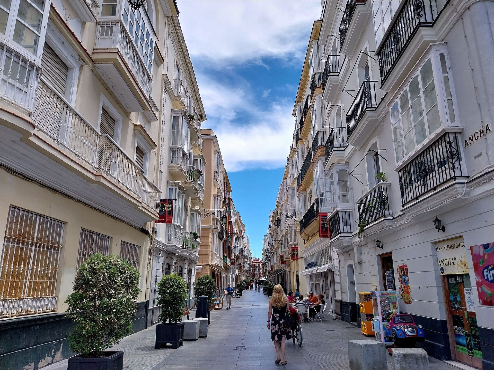 Ampia veduta di Calle Ancha a Cádiz con edifici storici, alberi in vaso, balconi aperti, caffè all'aperto e persone che passeggiano sotto un cielo luminoso.