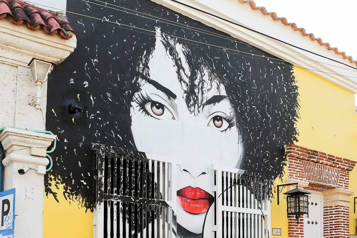 Large mural of a woman's face with curly hair and red lips on a yellow building along Calle del Arsenal in Getsemaní.