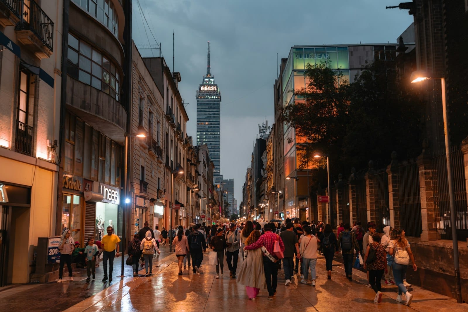 Vista serale di Calle Madero affollata di persone, con edifici coloniali che fiancheggiano la strada e la Torre Latinoamericana illuminata sullo sfondo.
