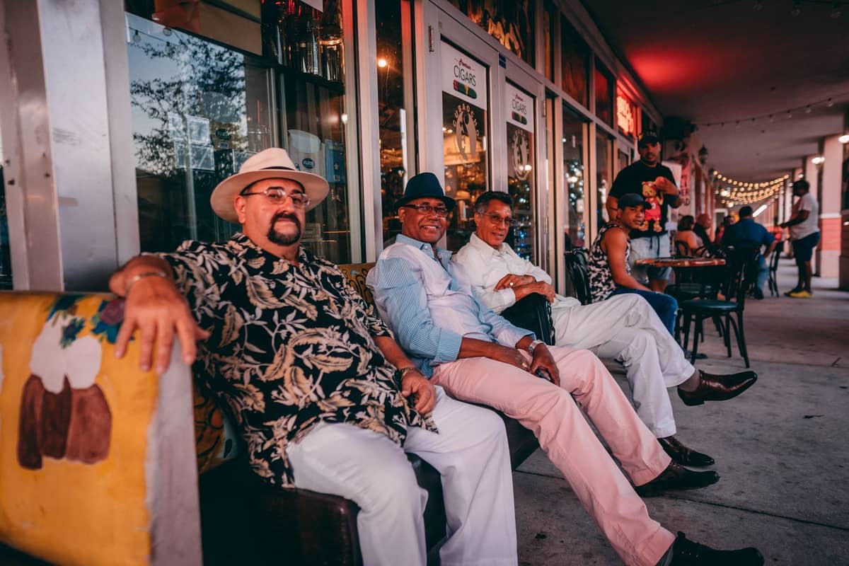 Group of people sitting and socializing on the sidewalk of a vibrant Miami street lined with cafes and string lights.