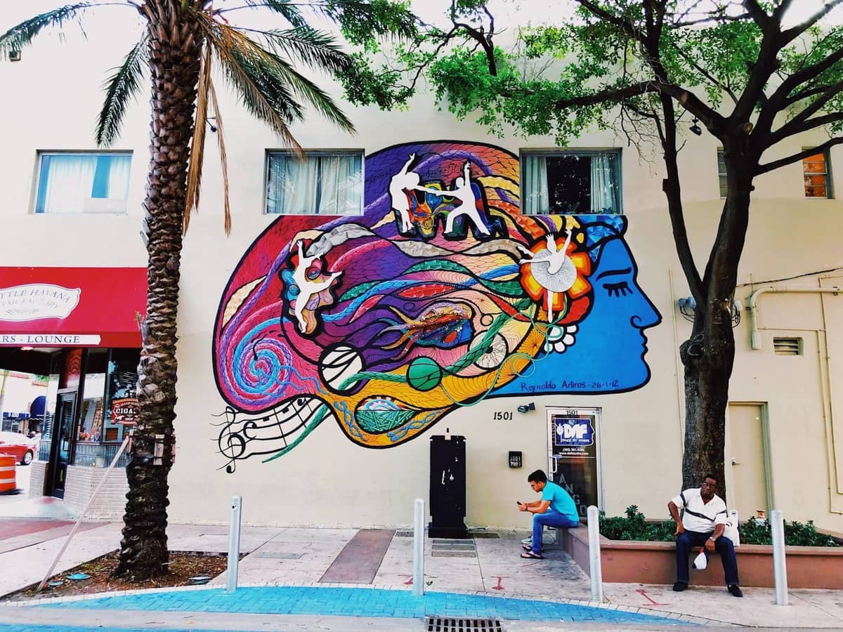 Street scene in Miami's Little Havana with colorful mural, palm tree, and people sitting by the sidewalk.
