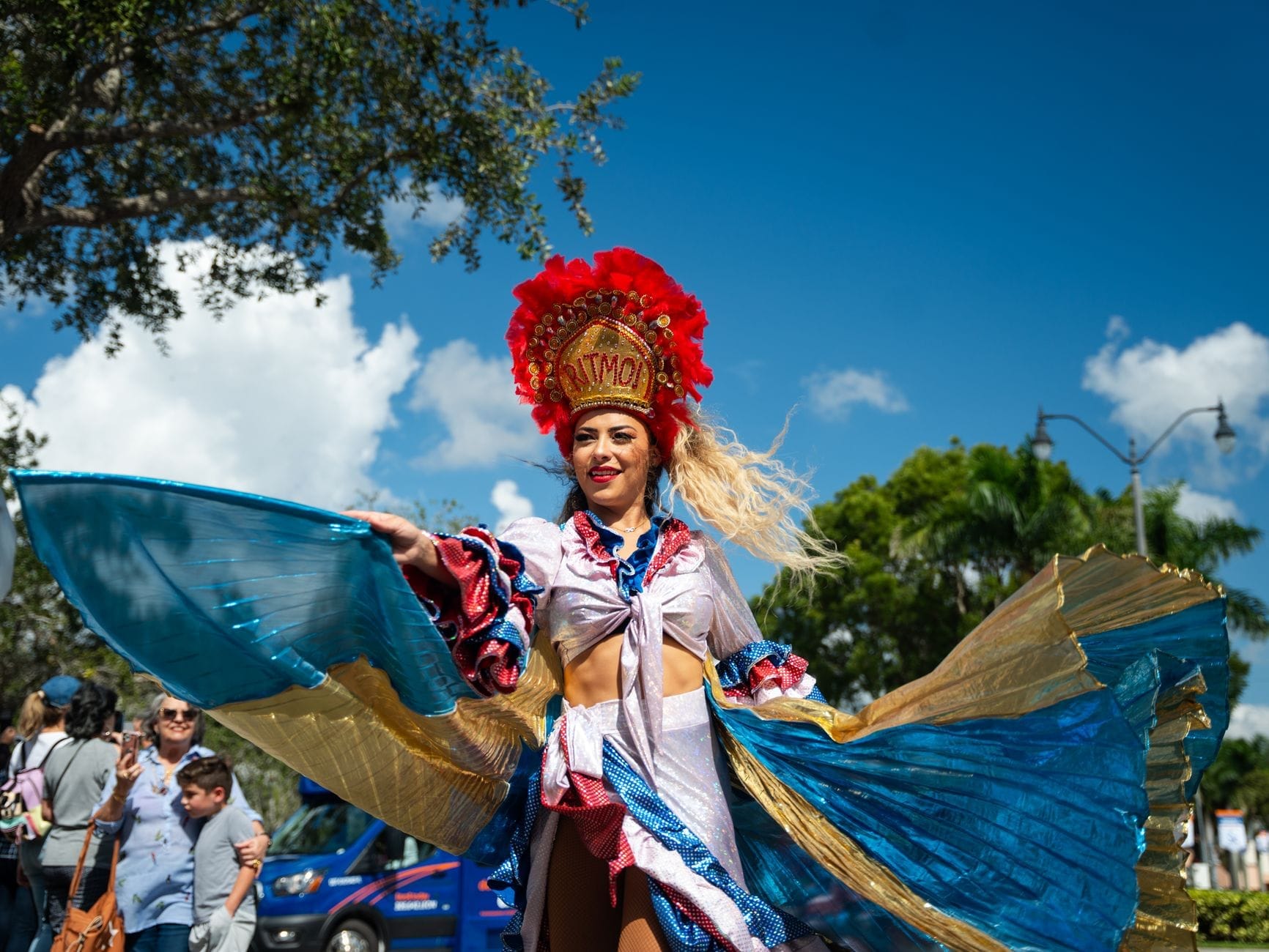 Mulher com fantasia carnavalesca elaborada e colorida, com cocar de penas, dança ao ar livre durante um festival de rua sob um céu azul intenso, com o público ao redor.