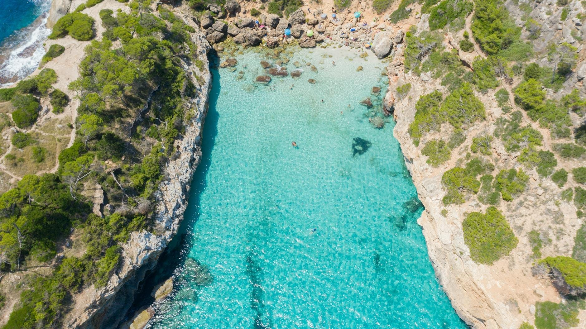 Vista aerea della stretta cala turchese di Caló des Moro, circondata da scogliere imponenti e pini, con una piccola spiaggia in fondo e un'acqua cristallina.