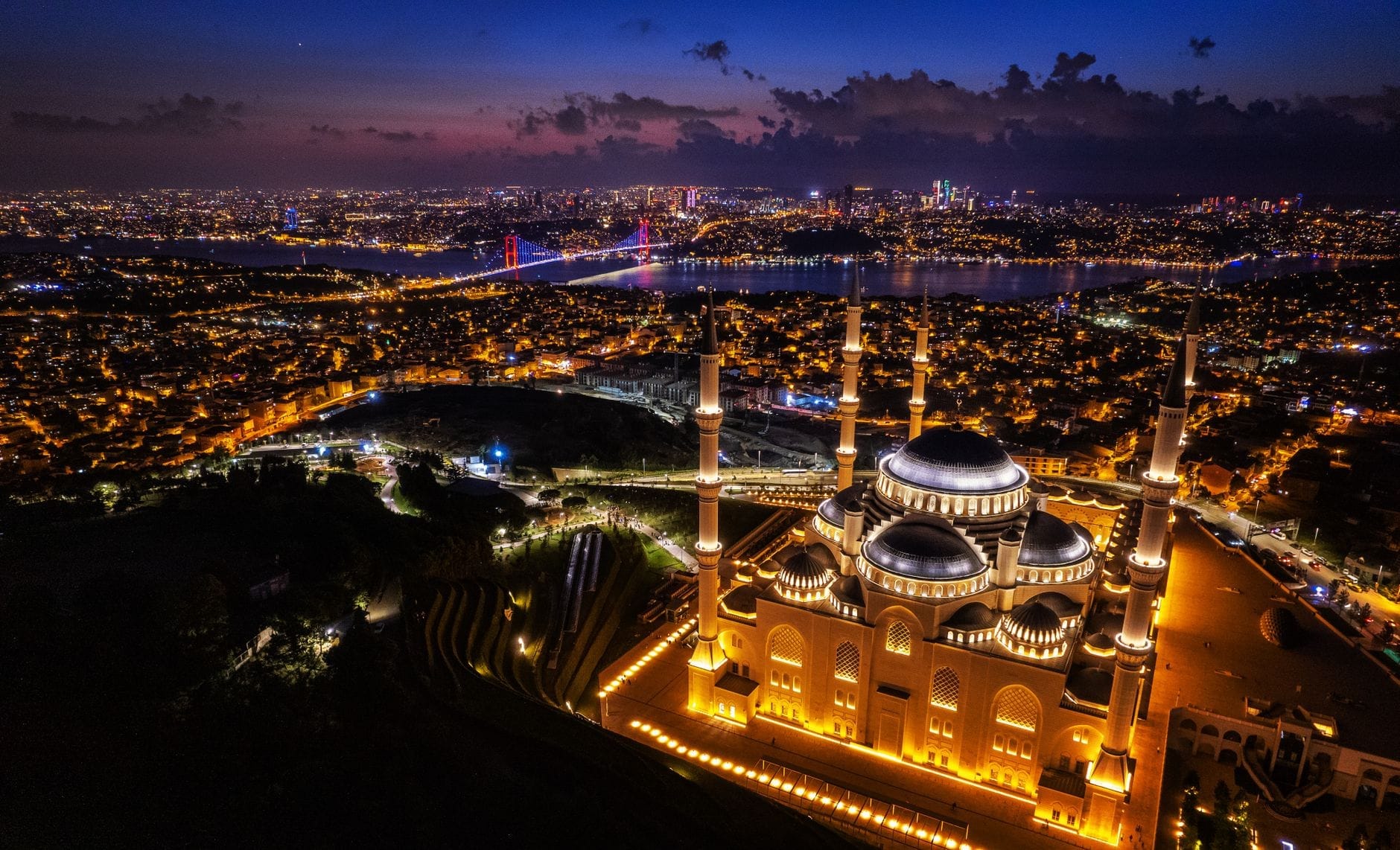 Vista aerea della Moschea di Çamlıca illuminata al tramonto, con lo skyline di Istanbul e il Ponte sul Bosforo sullo sfondo.