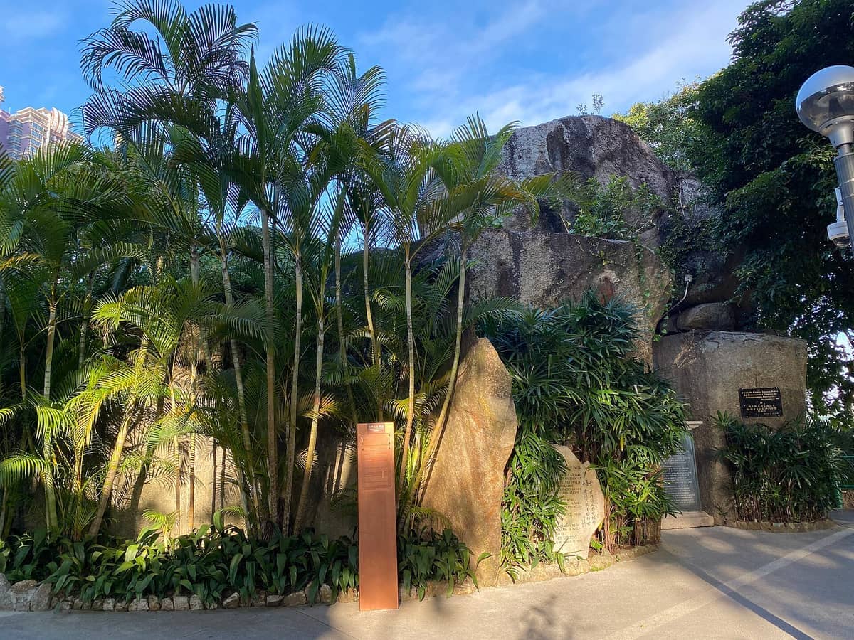 Hohe Palmen und große Felsformationen am Eingang zur Camões-Grotte in Macao, mit Informationstafeln und üppigem Grün.