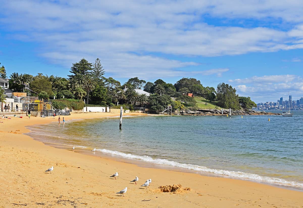 La playa de Camp Cove en Watsons Bay, con arena dorada, olas suaves y el horizonte de la ciudad visible a lo lejos en un día despejado.