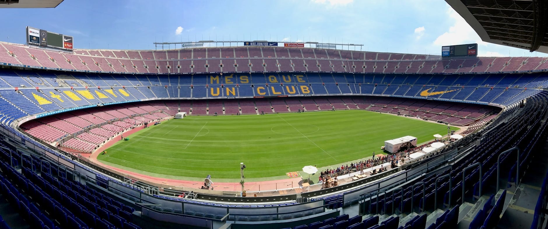 Wide panoramic view of the empty Camp Nou stadium from the stands with clear skies, showing the field and iconic MES QUE UN CLUB seats.