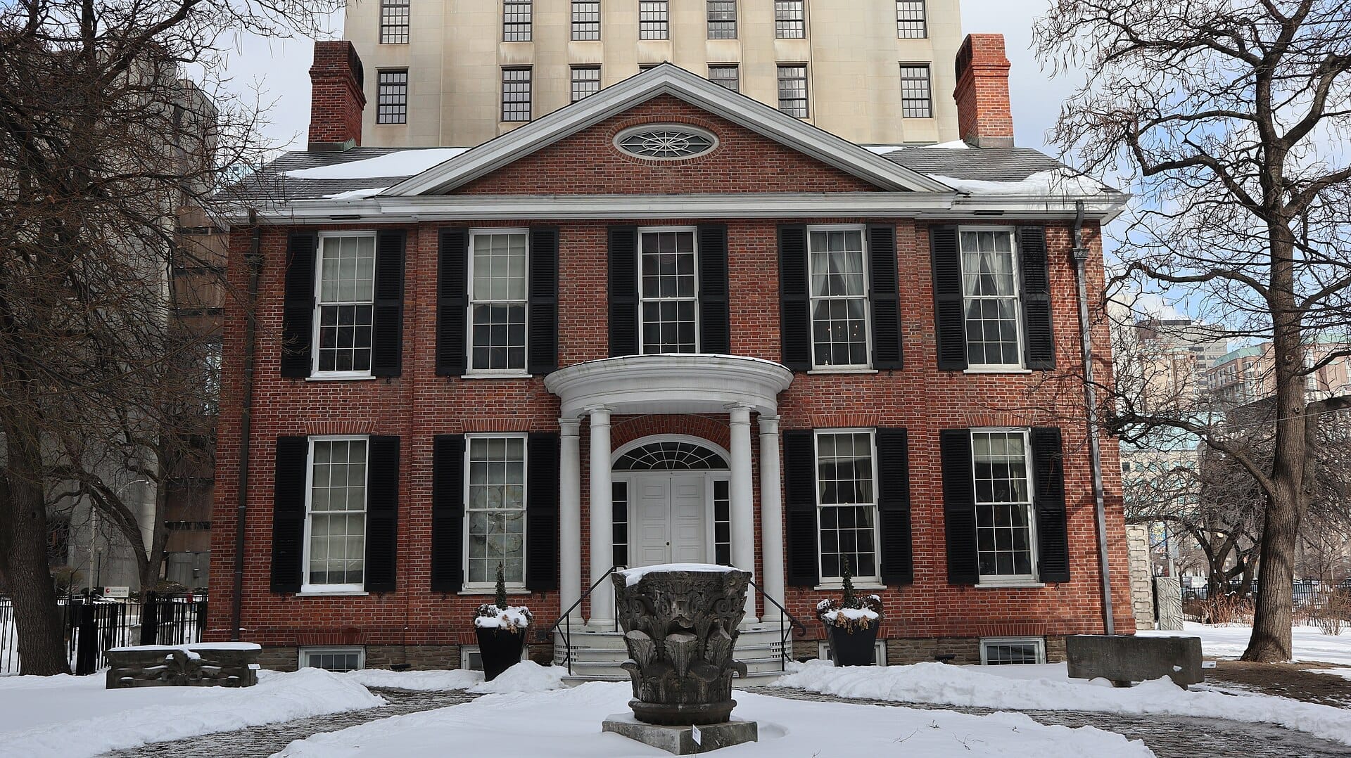 Campbell House Museum อาคารสไตล์จอร์เจียนก่ออิฐสีแดงพร้อมบานเกล็ดสีดำ ตั้งอยู่ท่ามกลางหิมะโดยมีอาคารขนาดใหญ่ในย่านดาวน์ทาวน์อยู่เบื้องหลัง
