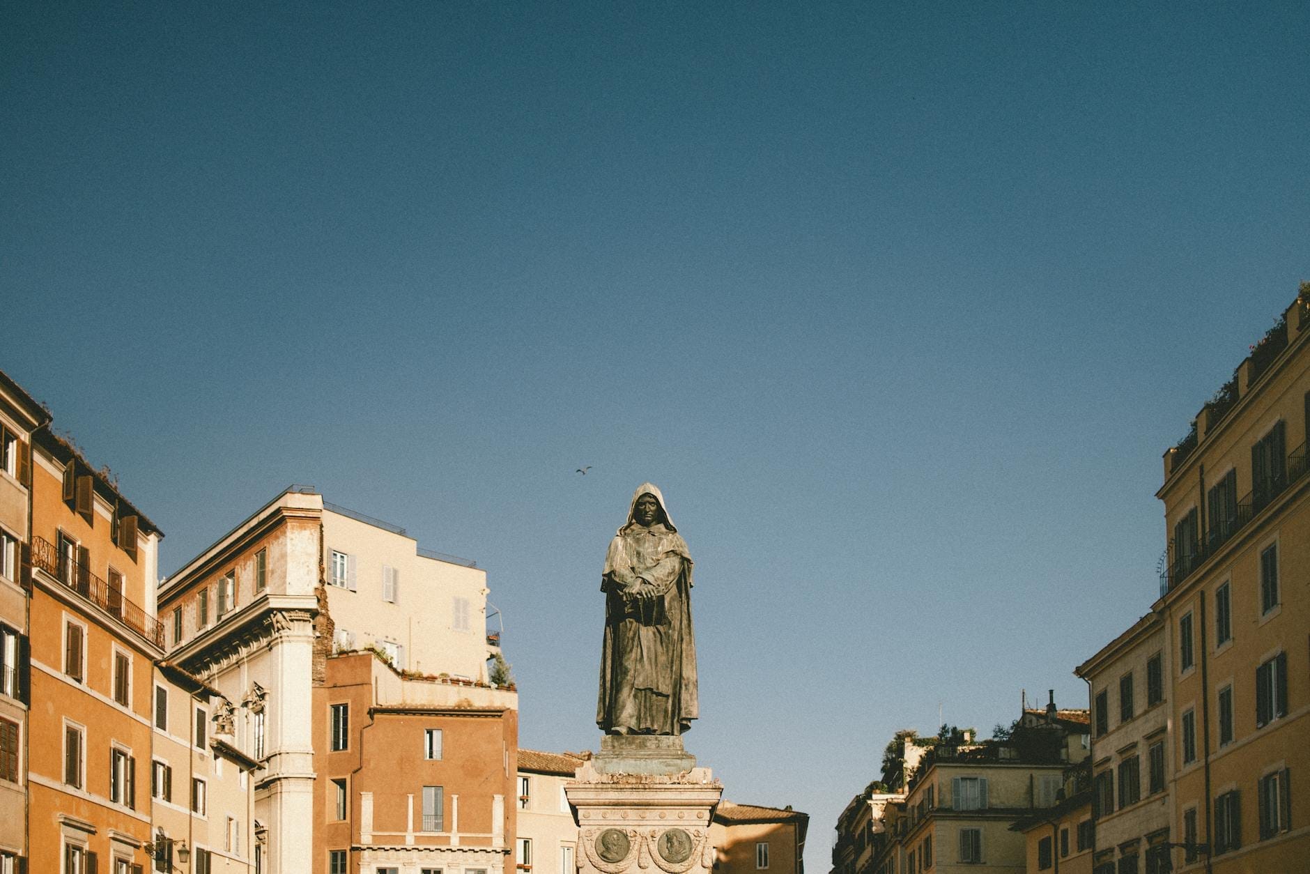 Statue of Giordano Bruno at the center of Campo de' Fiori square in Rome, surrounded by historic buildings and a clear blue sky.