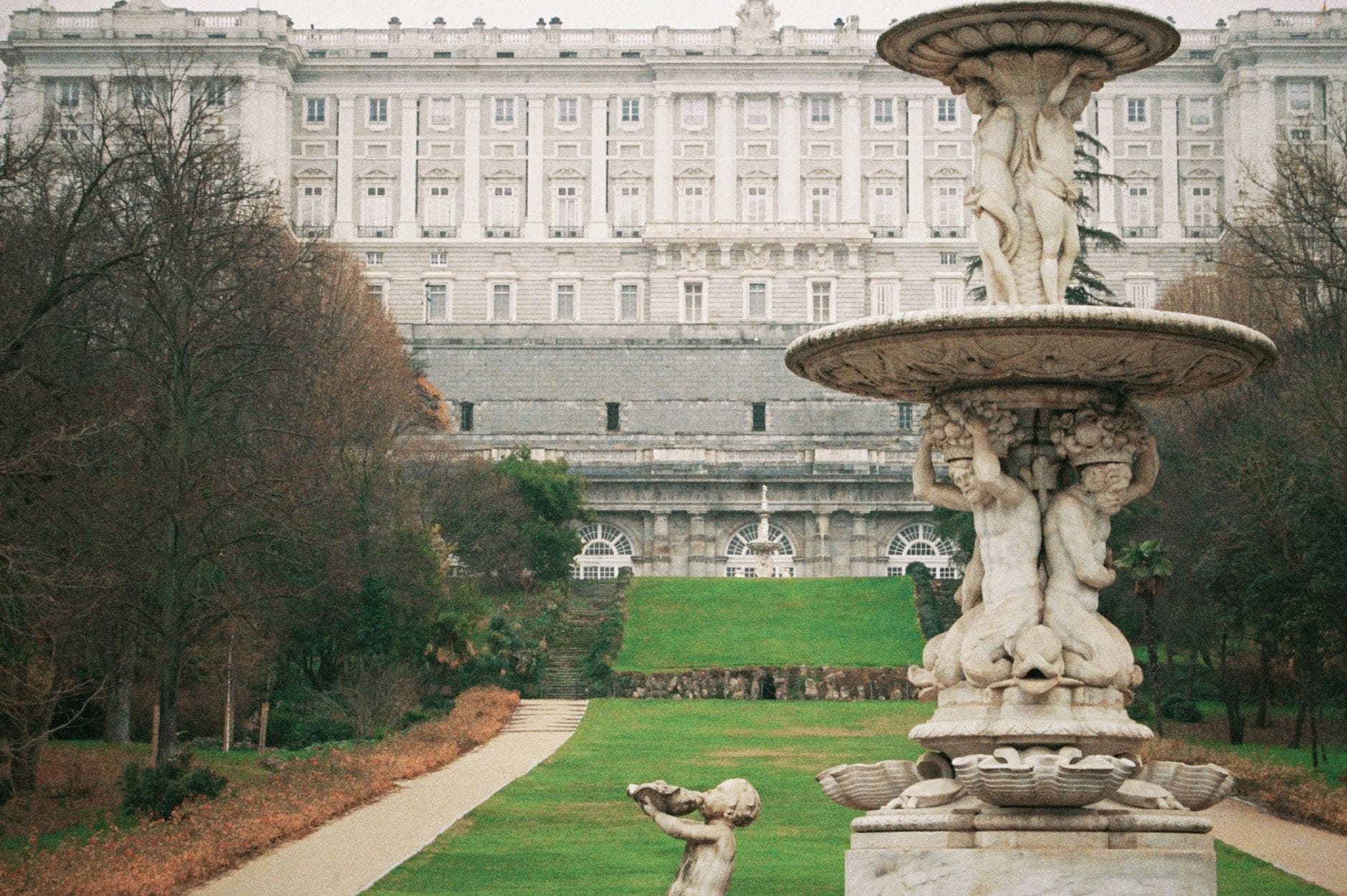 Vista del Palazzo Reale sullo sfondo di una grande fontana decorativa e dei vialetti curati dei Giardini del Campo del Moro in una giornata nuvolosa.