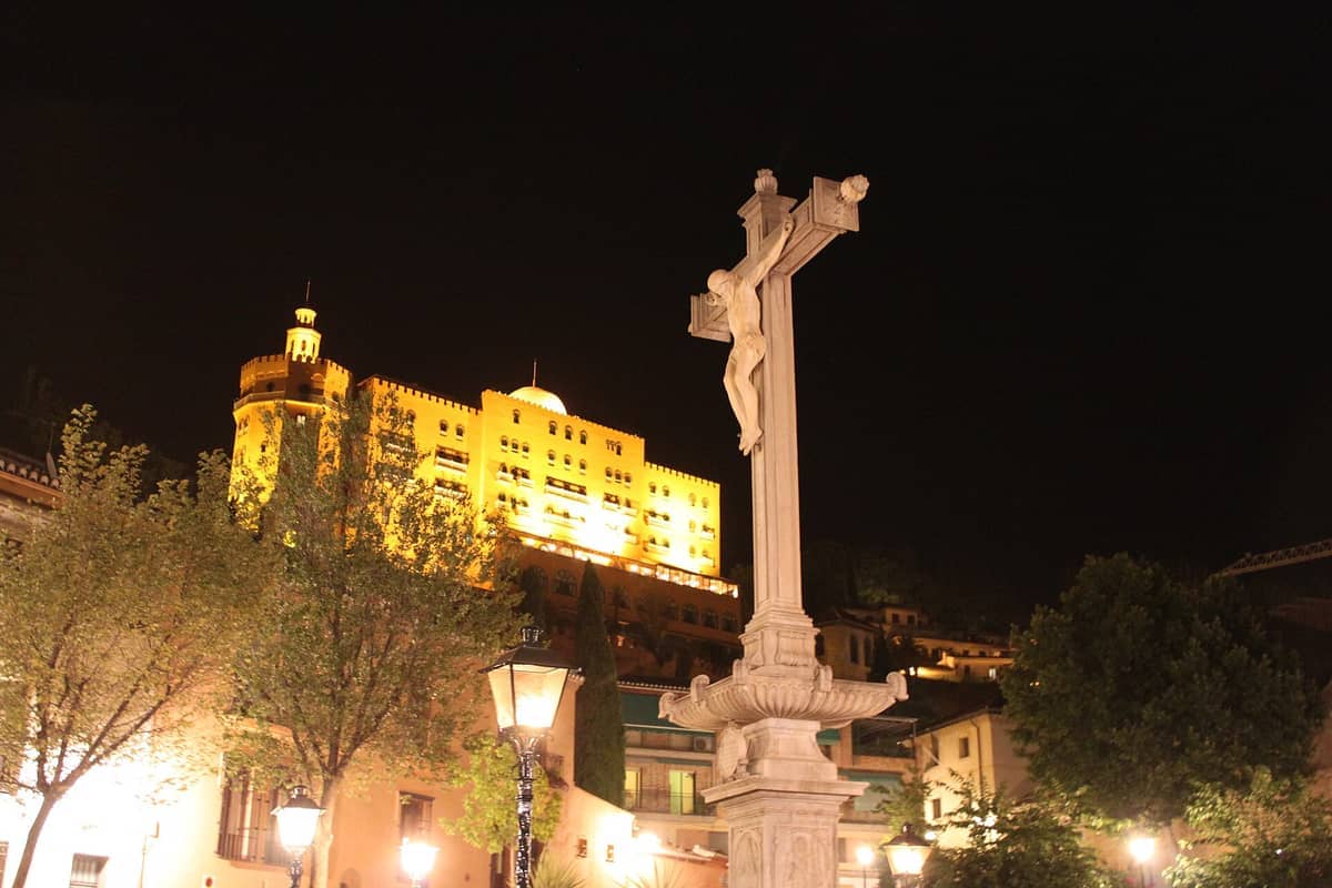 วิวยามค่ำคืนของ Campo del Príncipe ในเมือง Granada โดดเด่นด้วยรูปปั้นไม้กางเขนที่สว่างไสว ต้นไม้ใบหนาทึบ และอาคารทางประวัติศาสตร์โดยรอบ