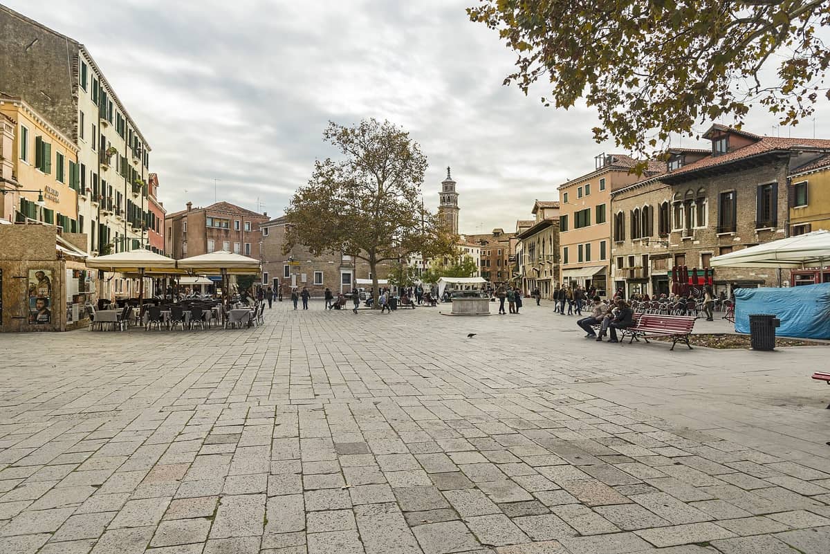 Weitwinkelansicht des Campo Santa Margherita in Venedig mit offenem Platz, Cafés, Bänken, Spaziergängern und entspannten Menschen sowie den Bäumen und markanten Gebäuden des Platzes im Hintergrund.