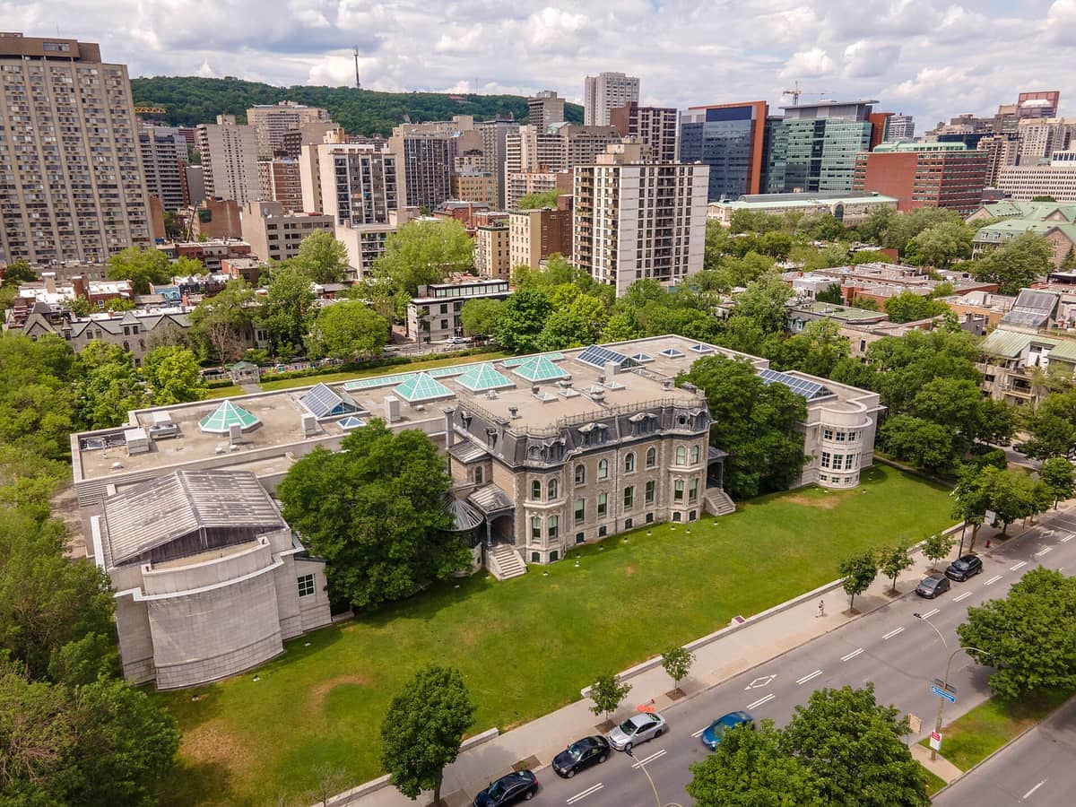 Luftaufnahme des Canadian Centre for Architecture mit seinem historischen Gebäude, umgeben von Bäumen, vor der modernen Stadtkulisse von Montreal.