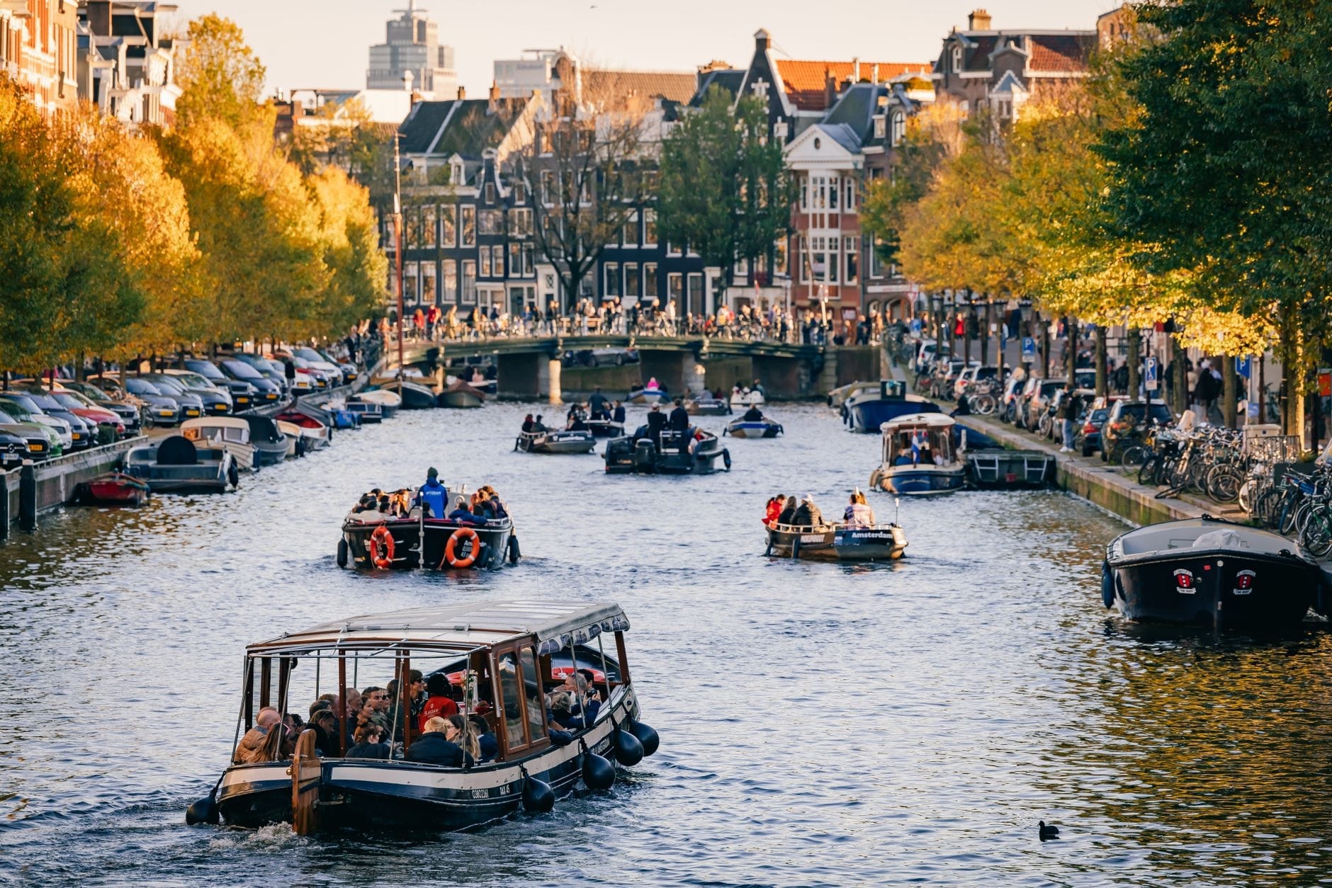 Varios barcos turísticos con pasajeros navegan por un canal de Ámsterdam flanqueado de árboles y edificios históricos en una tarde soleada.