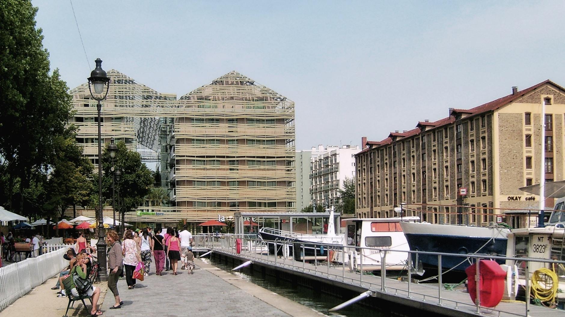Persone che passeggiano e si rilassano lungo il Canal de l'Ourcq, con barche ormeggiate e edifici moderni lungo il vivace lungocanale in una giornata di sole.