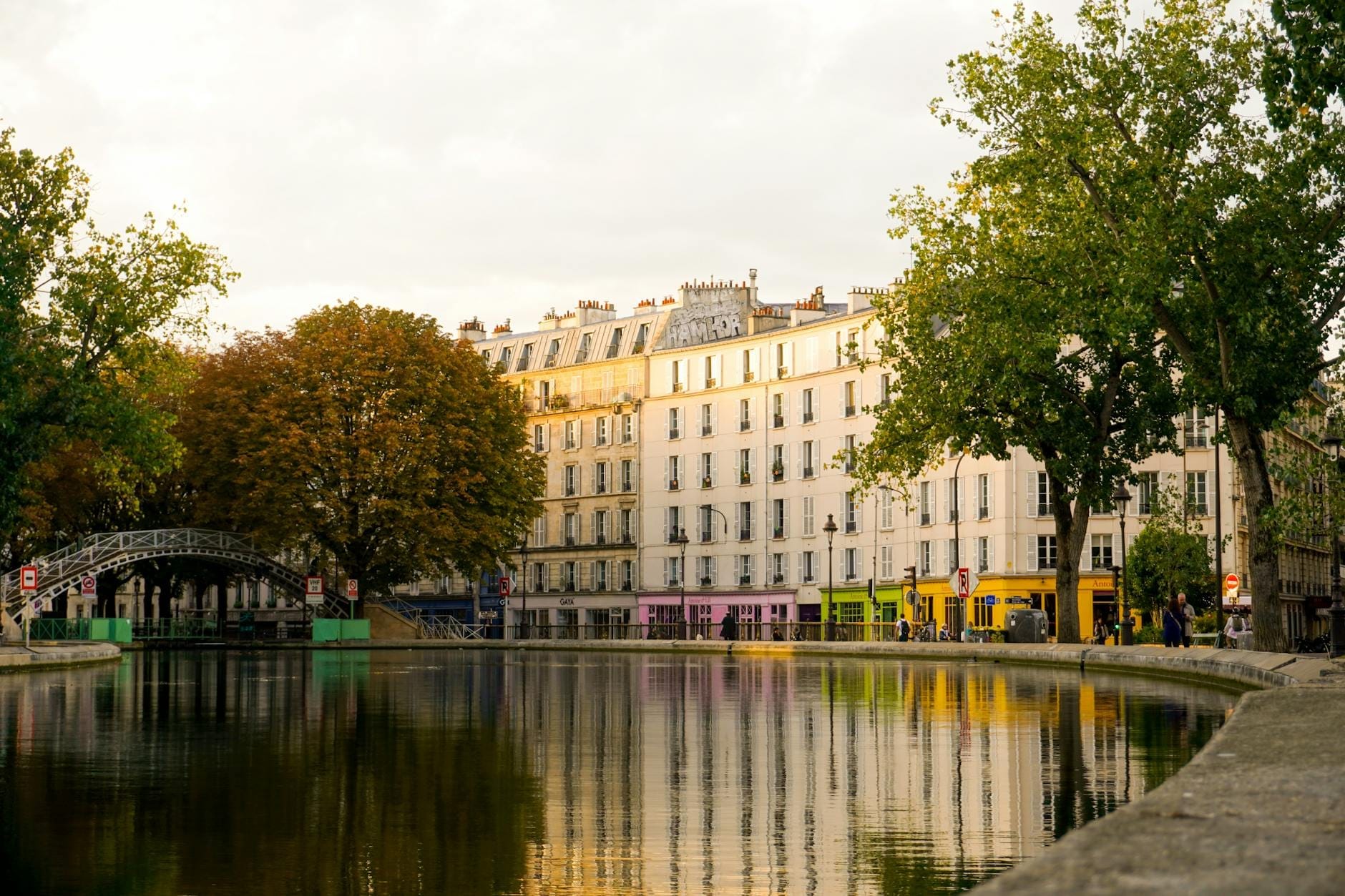 Un canale tranquillo fiancheggiato da alberi e palazzi parigini, negozi dai colori vivaci e una passerella in ferro ad arco che si riflette nell'acqua al tramonto.