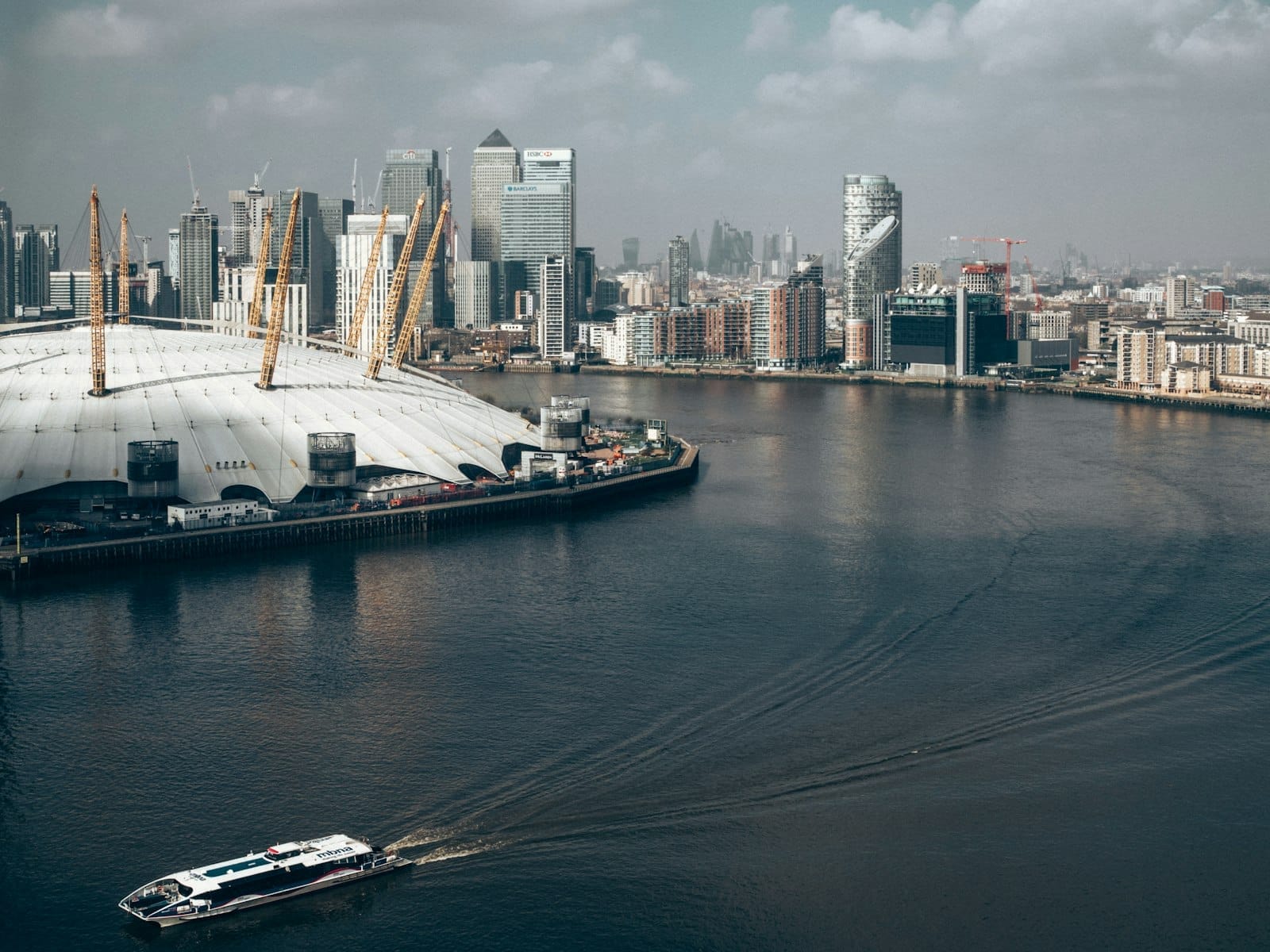 ภาพมองข้ามแม่น้ำเทมส์ไปทาง O2 Arena และสกายไลน์ของ Canary Wharf มีอาคารระฟ้าทันสมัย ริมน้ำ และเรือแล่นอยู่ใต้ท้องฟ้าครึ้มเมฆ