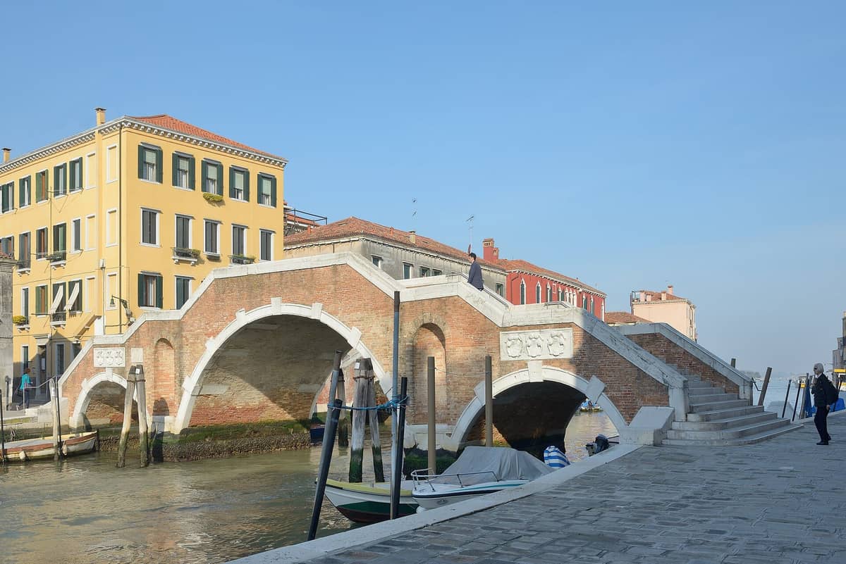 Panoramablick auf den Ponte dei Tre Archi, die ikonische Brücke mit drei Bögen über den Cannaregio-Kanal in Venedig, mit bunten Gebäuden und Menschen in der Nähe.