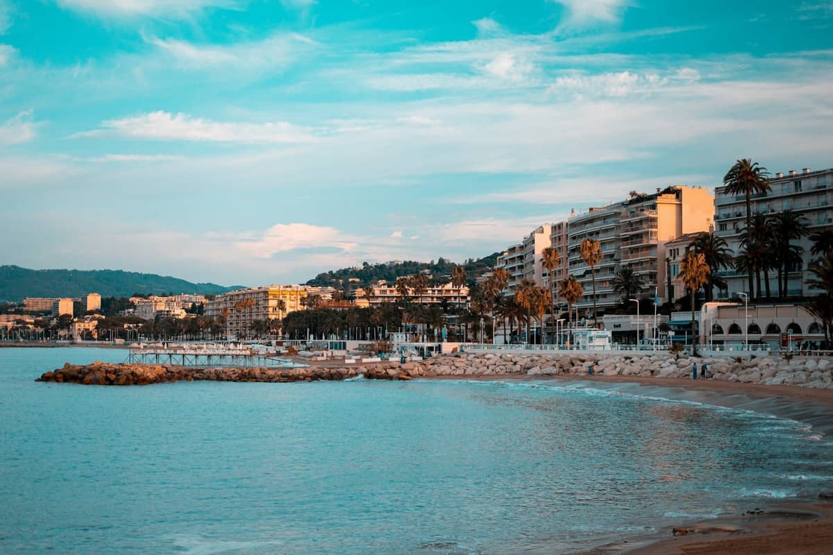 Vista della spiaggia di Cannes con acqua blu, riva sabbiosa, palme e grandi hotel lungo il lungomare sotto un cielo vivace.