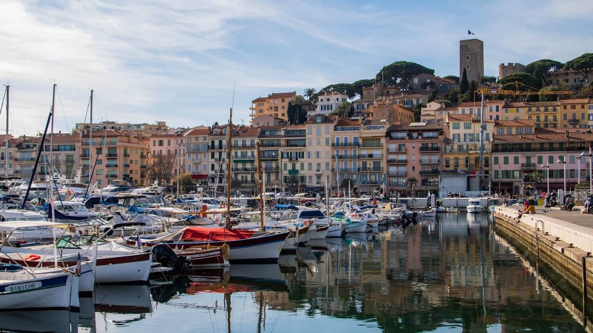 Vista del Vieux Port di Cannes con barche, edifici sul lungomare e la torre storica di Le Suquet sullo sfondo in una giornata di sole.