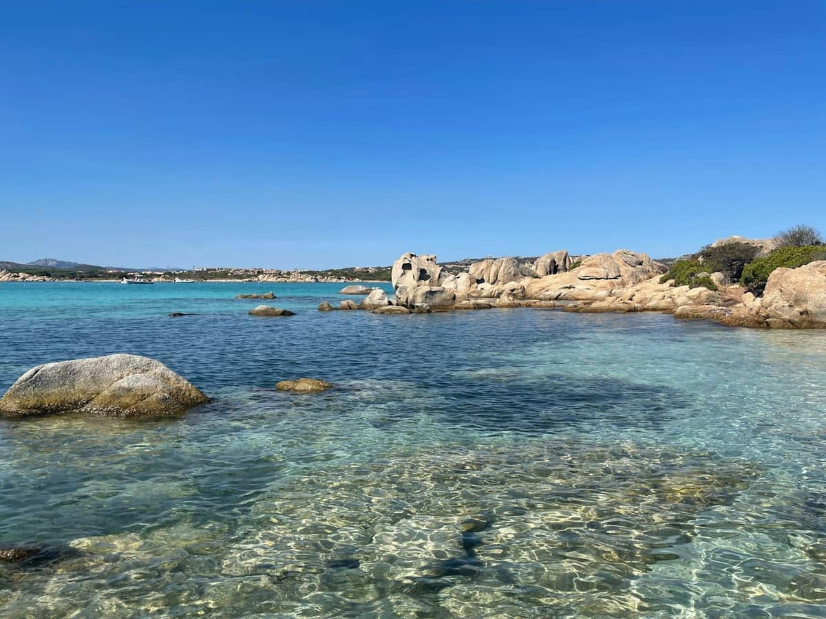 Acqua turchese limpida e poco profonda con rocce sparse e una costa naturale e accidentata sotto un cielo azzurro brillante: affascinante scena di spiaggia su un'isola nascosta.