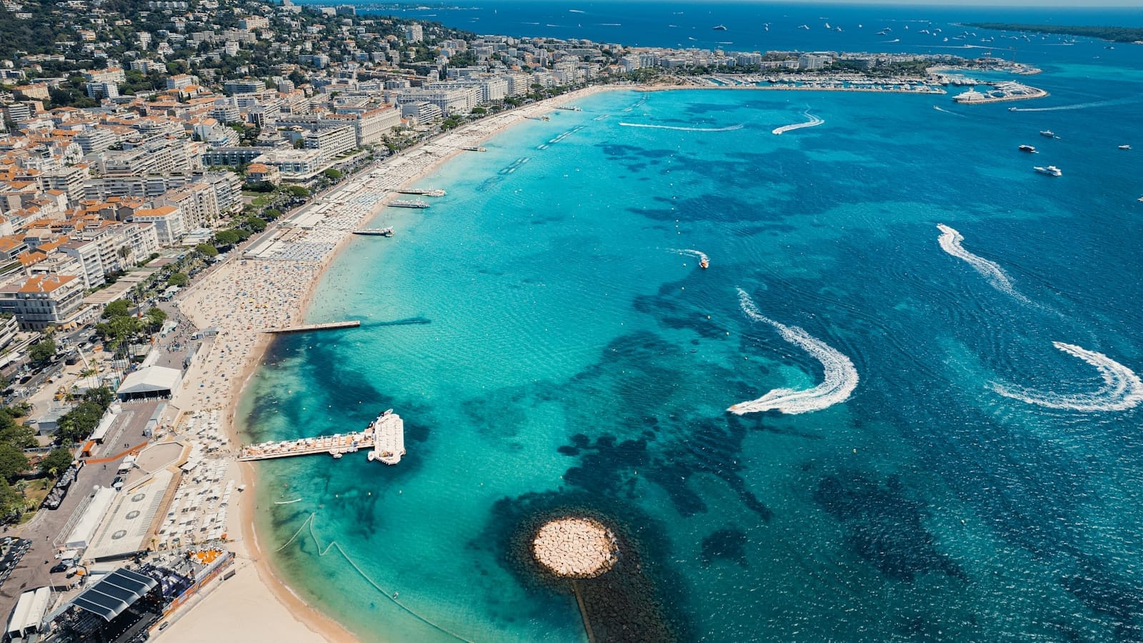 Vista aérea panorâmica de Cannes mostrando águas turquesa, praias de areia, clubes de praia e a orla da cidade sob um céu azul limpo.