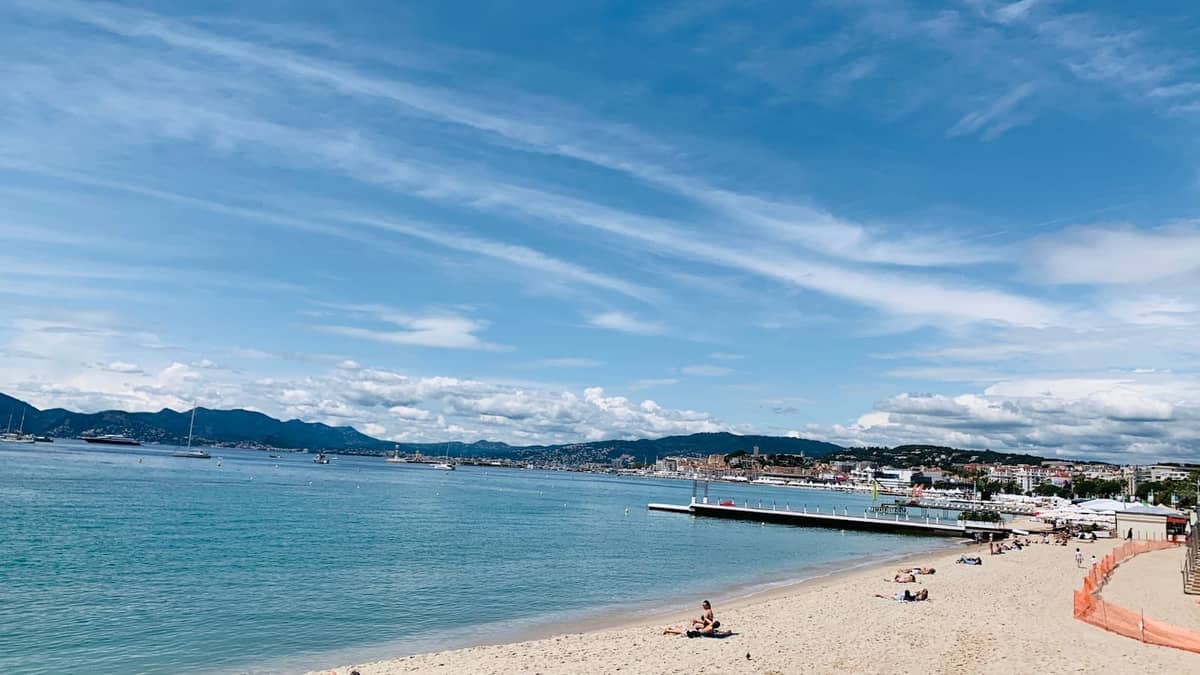 Ampia spiaggia pubblica sabbiosa a Cannes con mare blu calmo, bagnanti sparsi e vista lontana della città e delle montagne sotto un cielo soleggiato.