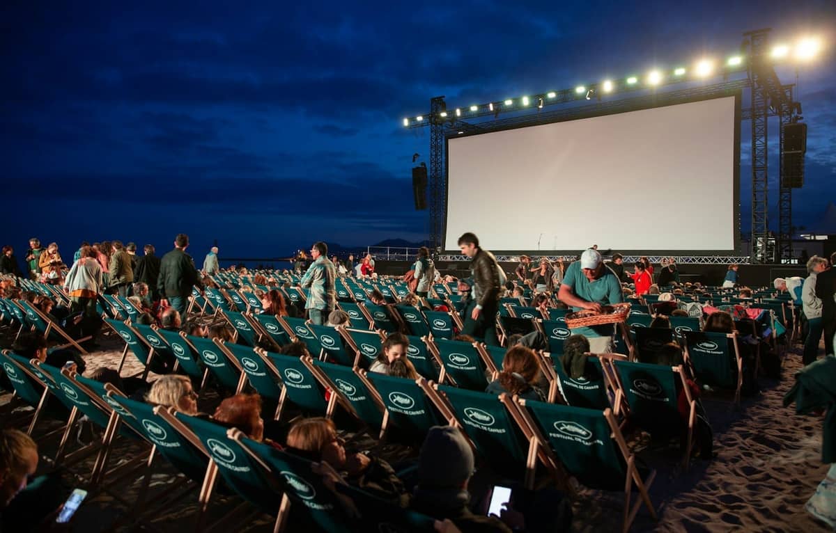Outdoor evening beach cinema with rows of people seated in chairs watching a large screen during the Cannes Film Festival.