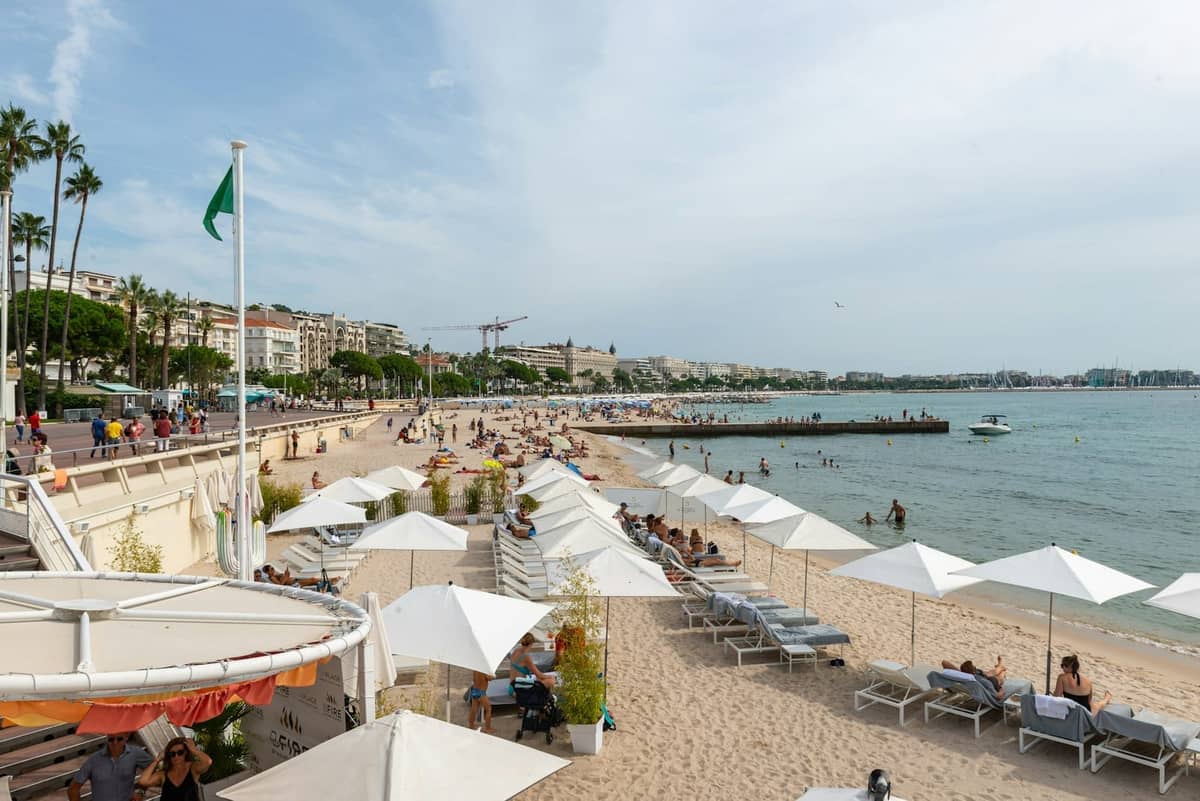 Scena na plaży w Cannes z rzędami leżaków i parasoli, odpoczywającymi turystami oraz miastem i linią brzegową w tle.