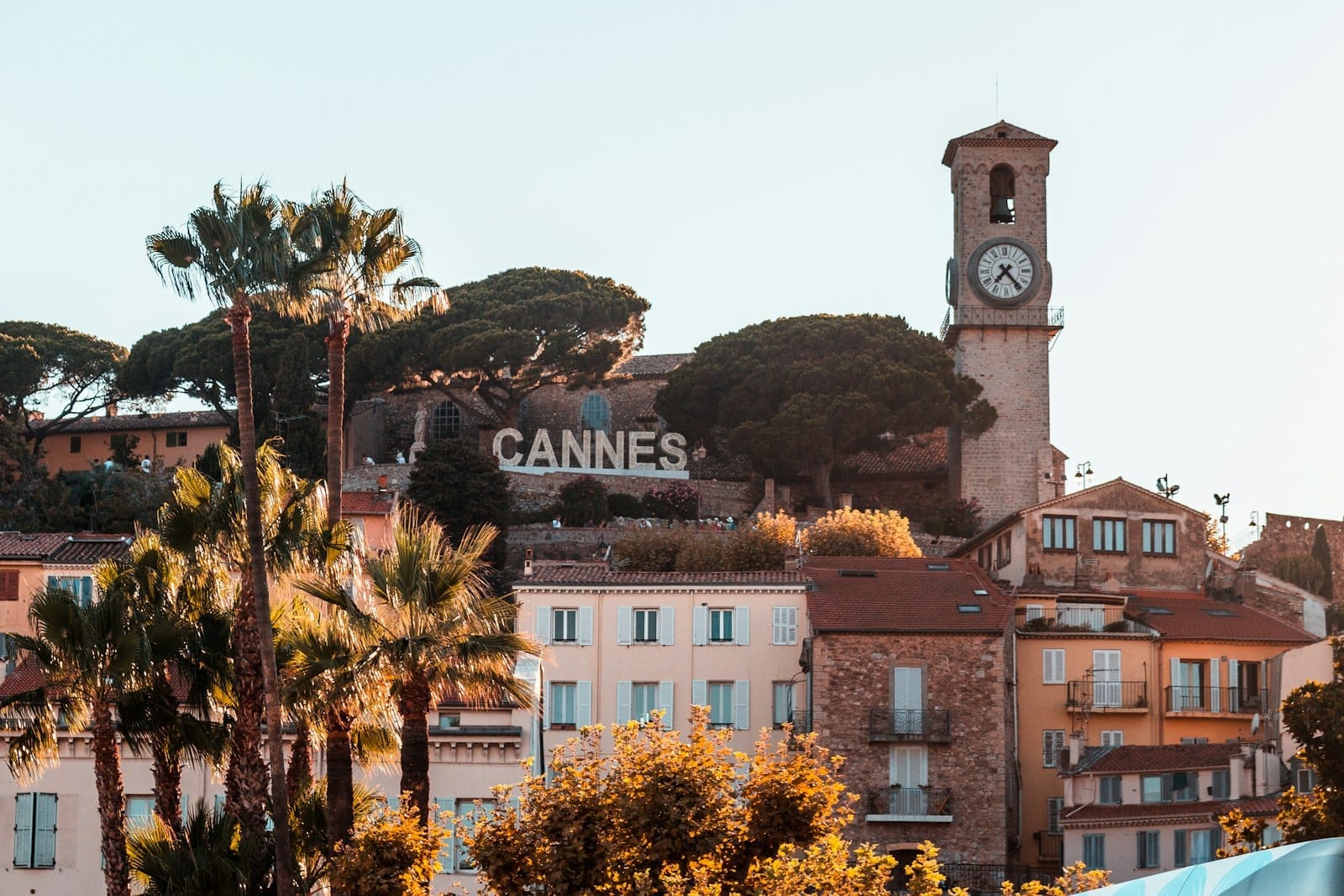 Vista do centro histórico de Cannes com prédios em tons pastéis, palmeiras, a icônica torre do relógio e o letreiro de Cannes na encosta ao pôr do sol.