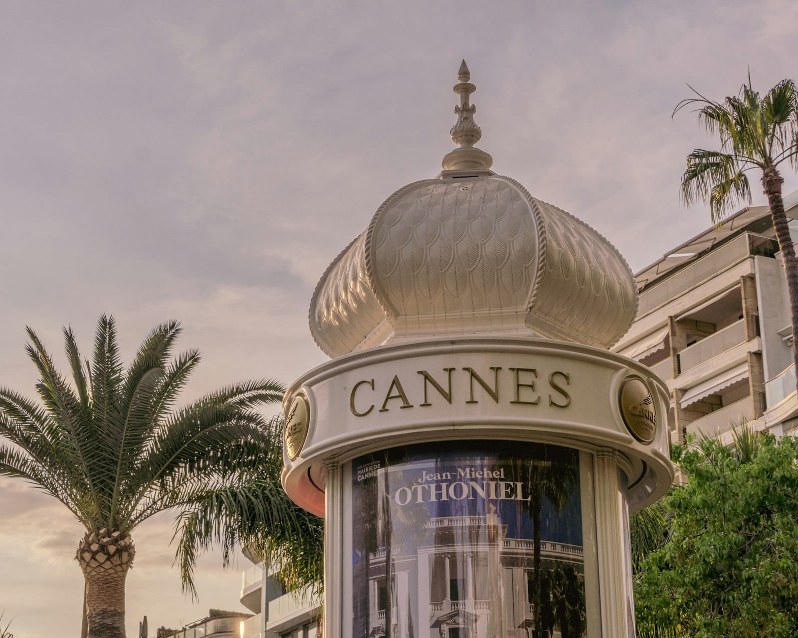 Quiosque em Cannes com topo ornamentado em forma de coroa, palmeiras e arquitetura elegante ao fundo, evocando uma atmosfera chique e convidativa para as compras.