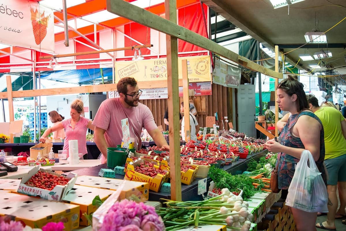 Escena de mercado interior con vendedores que ofrecen productos frescos, incluyendo fresas y verduras, a compradores bajo coloridos toldos.