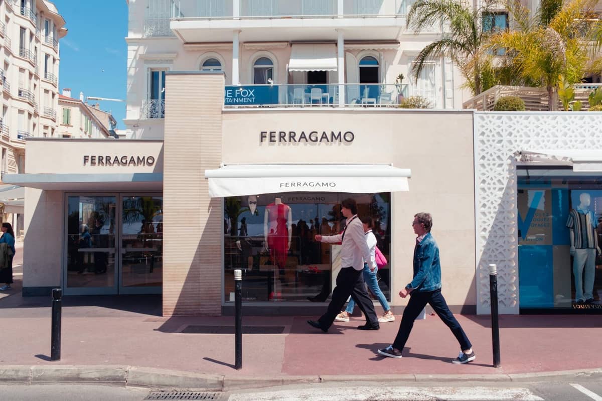 Personas pasan frente a la elegante boutique Ferragamo en el bulevar marítimo de Cannes, con elegantes edificios y palmeras al fondo.