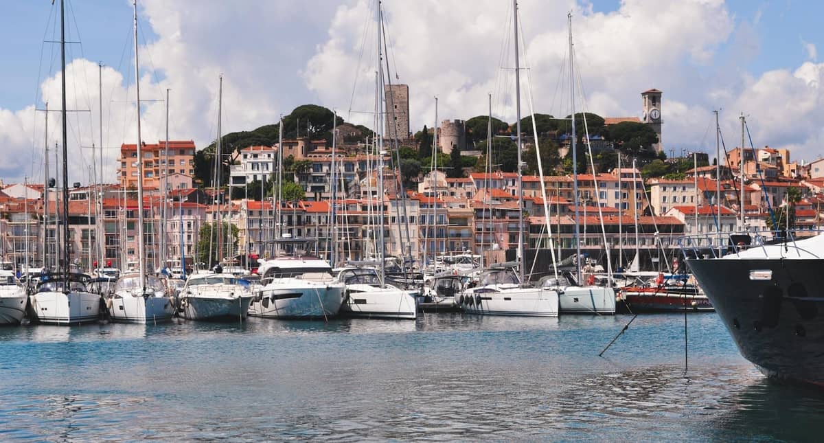 Cannes limanında demirlemiş yatlar ve arkasında yükselen tarihi şehir merkeziyle Cannes sahil şeridinin görünümü, kısmen bulutlu bir gökyüzü altında.