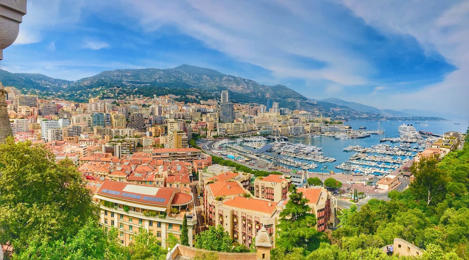 Vista panorâmica da paisagem urbana do Mónaco com edifícios coloridos, árvores verdejantes e um movimentado porto repleto de iates sob um céu azul radiante.