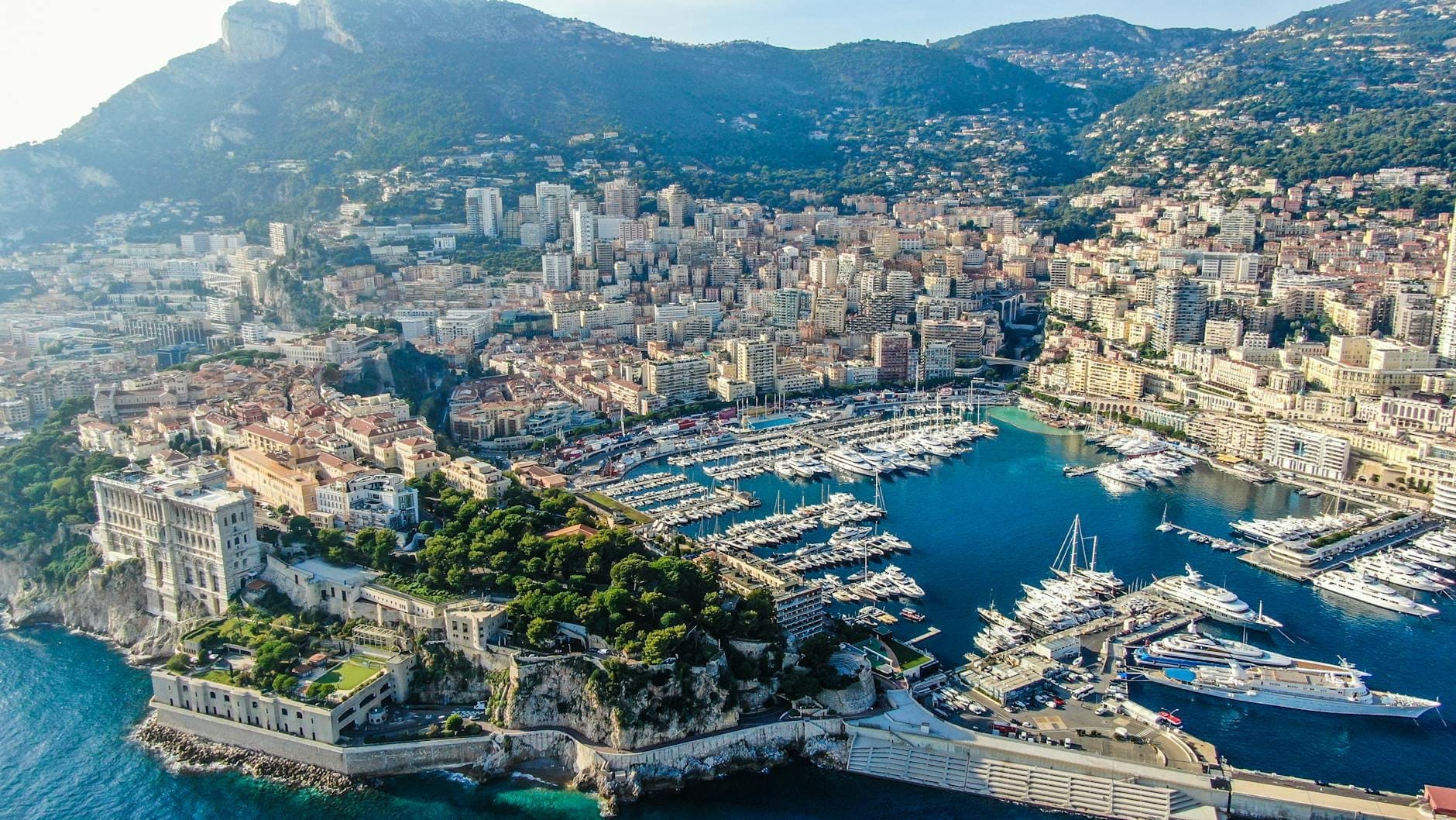 Vista aérea do porto de Mônaco com iates de luxo, edifícios históricos, colinas verdes e mar azul claro sob a luz do dia.
