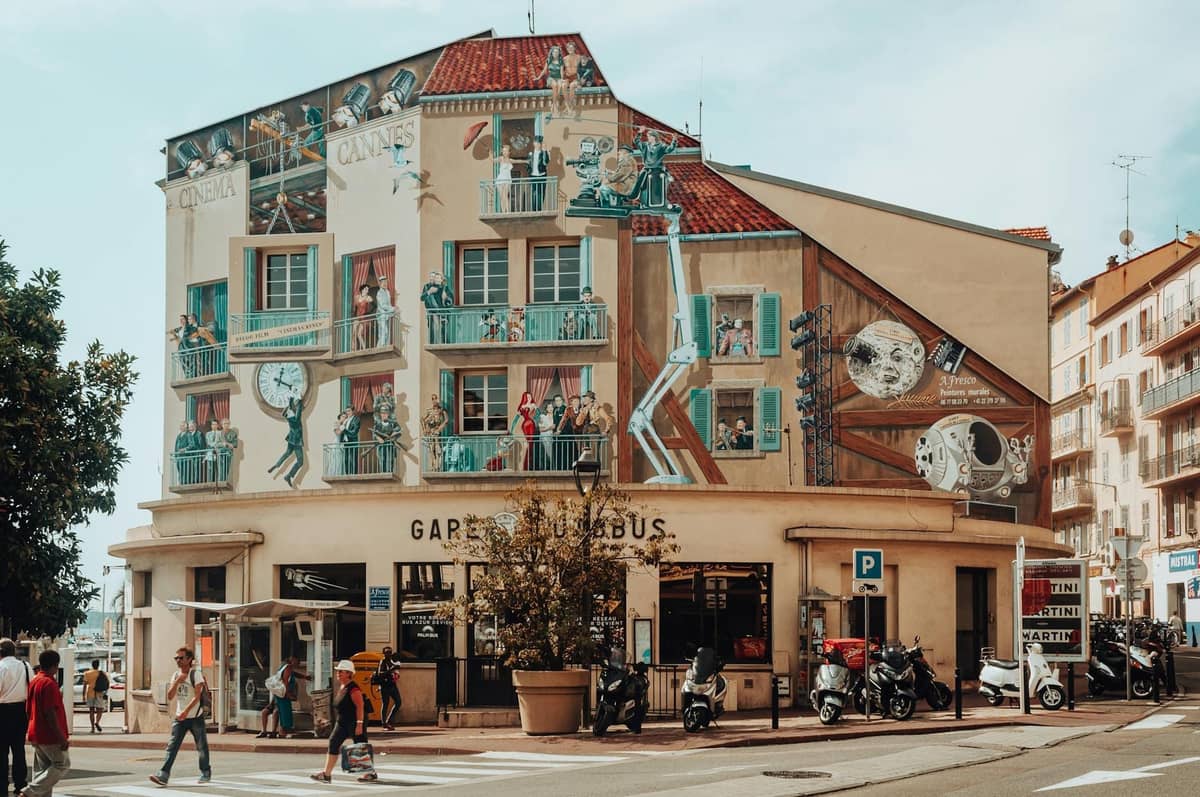 Ein Gebäude in Cannes, das mit bunten Wandmalereien geschmückt ist, die Filmszenen, Menschen und Balkone zeigen, lebhafte Straßenatmosphäre mit Fußgängern und Rollern.