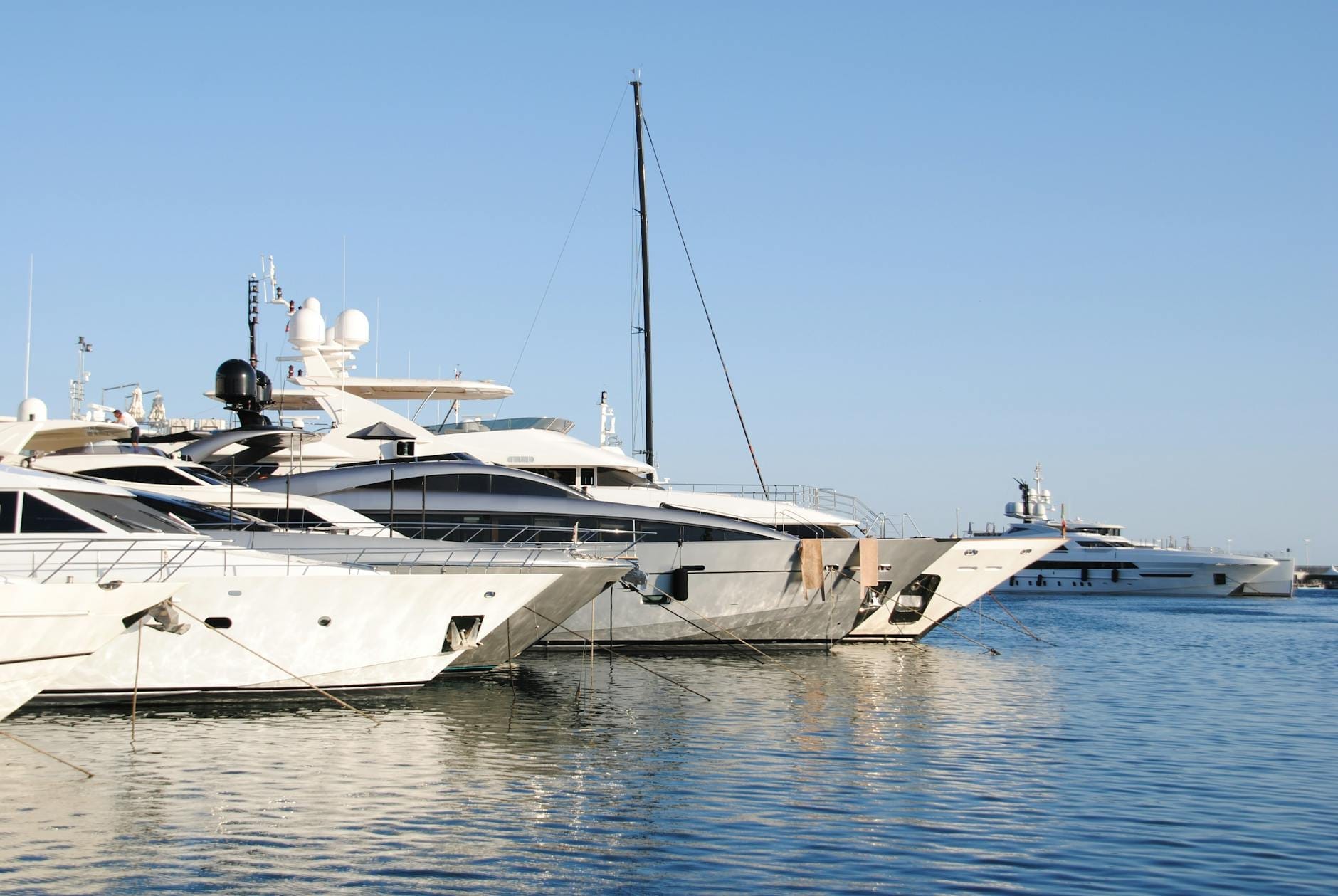 Fila di eleganti superyacht bianchi ormeggiati in una marina tranquilla, cielo blu e mare calmo che riflettono la luce per una scena di lusso sul lungomare.