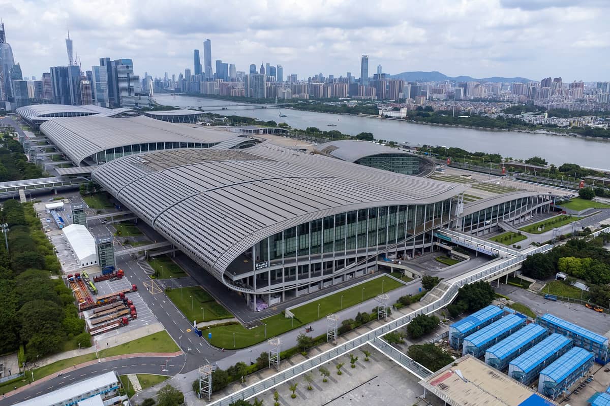 Aerial view of the massive Canton Fair Complex exhibition center in Guangzhou with its distinctive curved roof, seen beside the Pearl River and city skyline.