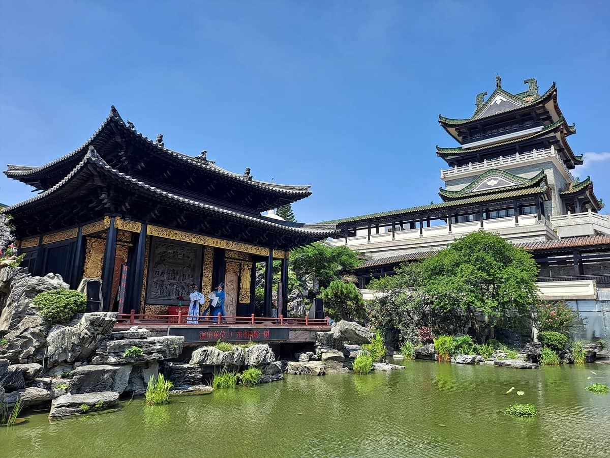 Traditional Lingnan architecture and a lush pond at Cantonese Opera Art Museum, with performers on an outdoor stage under sunny skies.