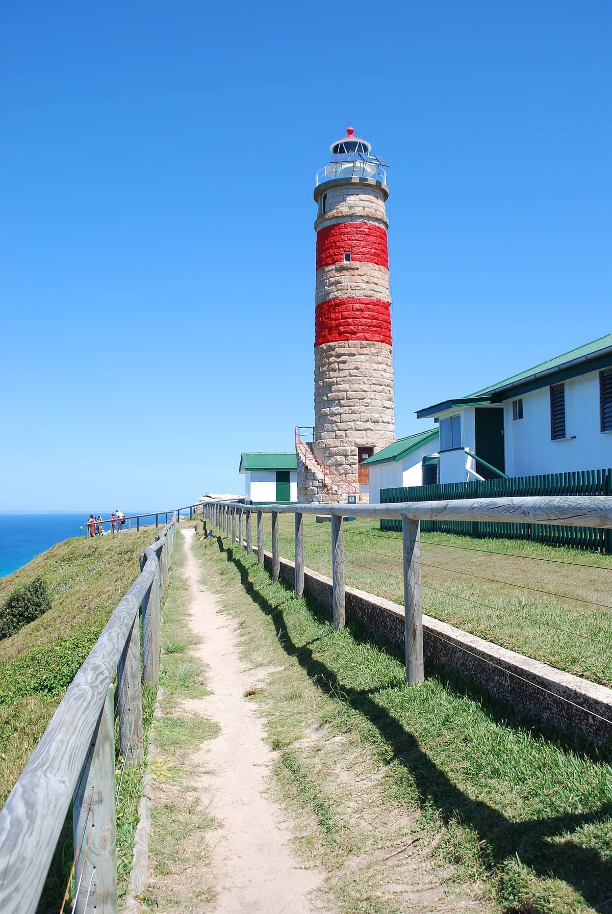 Der Cape Moreton Leuchtturm ragt mit roten und weißen Streifen in die Höhe, umgeben von Gebäuden mit grünen Dächern und einem Feldweg unter klarem blauem Himmel.
