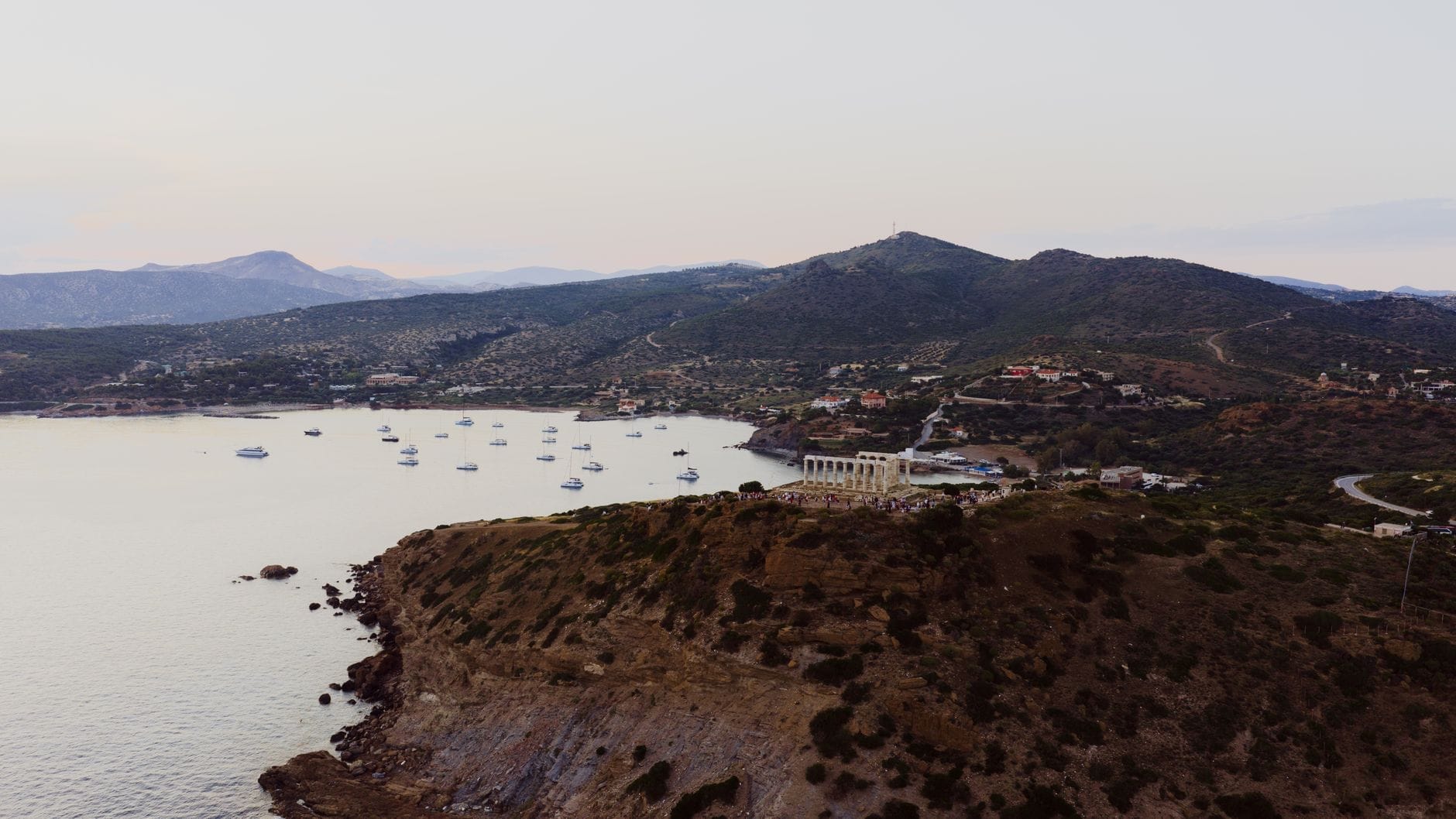 Una spettacolare vista aerea di Capo Sunio con il Tempio di Poseidone arroccato su una scogliera sopra il mare Egeo, con barche ancorate e dolci colline sullo sfondo.