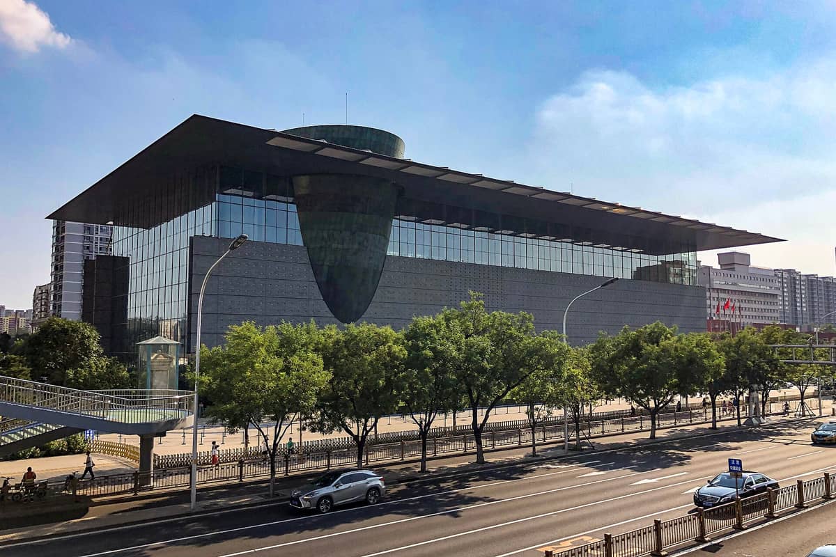 Außenansicht des modernen Capital Museums in Peking mit großen Glasfassaden und einem markanten bronzefarbenen Baukörper über einer belebten, baumgesäumten Straße.