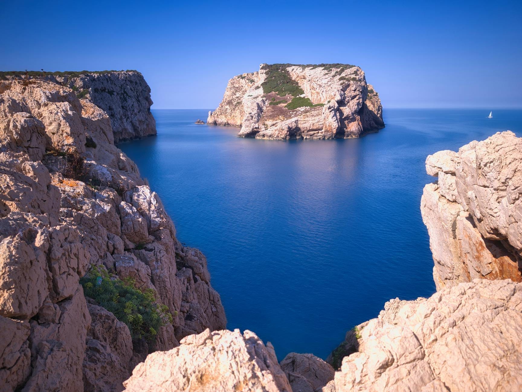 Dramatische Kalksteinklippen und das raue Vorgebirge von Capo Caccia, das sich über dem tiefen blauen Meer erhebt – aufgenommen von einem hoch gelegenen Felsvorsprung unter strahlend blauem Himmel.