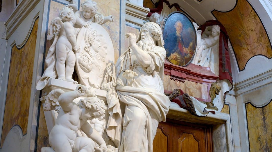Dettagliate sculture in marmo di figure e cherubini all'interno della Cappella Sansevero, con sfondi ornati e un dipinto ritratto sopra una porta in legno.