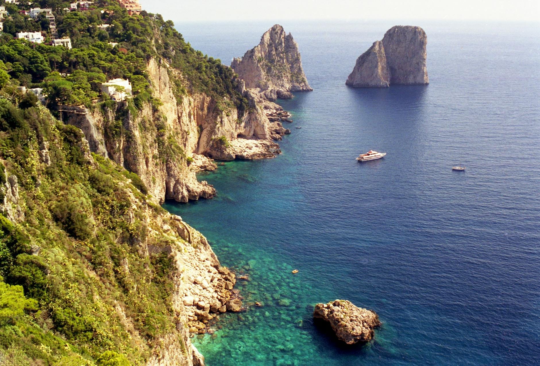 Le spettacolari falesie di calcare di Capri si ergono sopra le acque blu intenso del Mediterraneo, con gli iconici Faraglioni e le barche che punteggiano il mare sottostante.