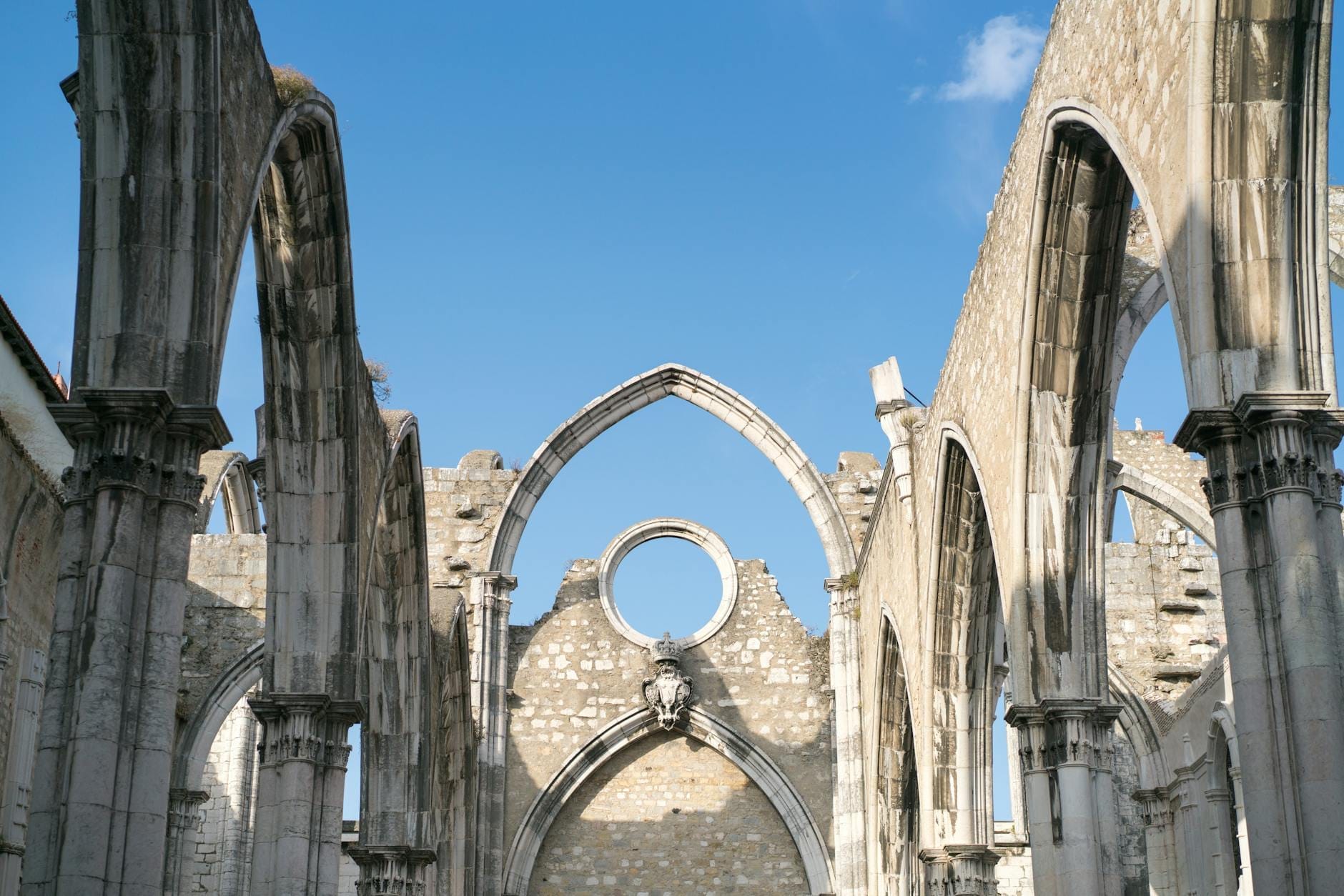 Gli archi gotici in rovina e le mura in pietra del Convento do Carmo a Lisbona, aperti verso un cielo azzurro intenso, evocano un senso di storia e atmosfera sospesa nel tempo.