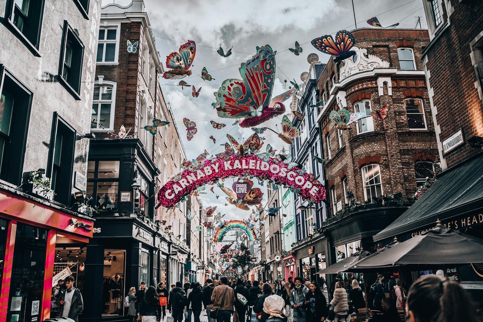 Carnaby Street a Londra affollata di persone, con negozi colorati, decorazioni a farfalla sospese e un arco festivo con la scritta 'Carnaby Kaleidoscope'.