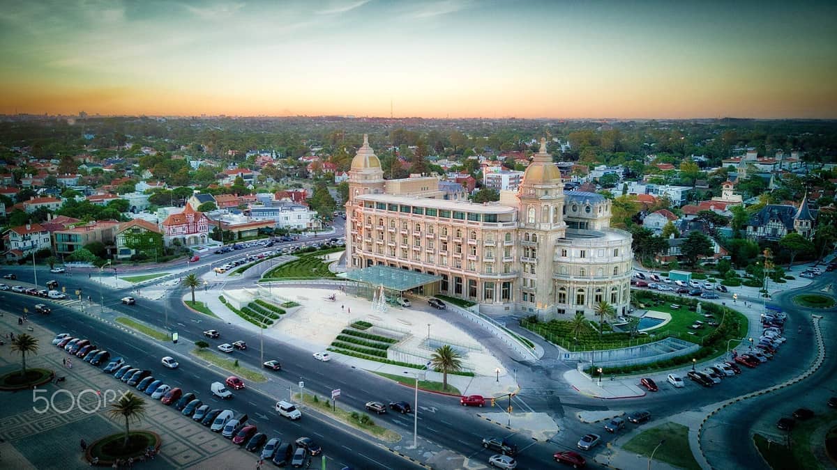 มุมมองทางอากาศของ Hotel Carrasco อันเก่าแก่ที่ล้อมรอบด้วยถนนเรียงรายต้นไม้และบ้านพักอาศัยในย่านหรู Carrasco ของมอนเตวิเดโอยามพระอาทิตย์ตกดิน