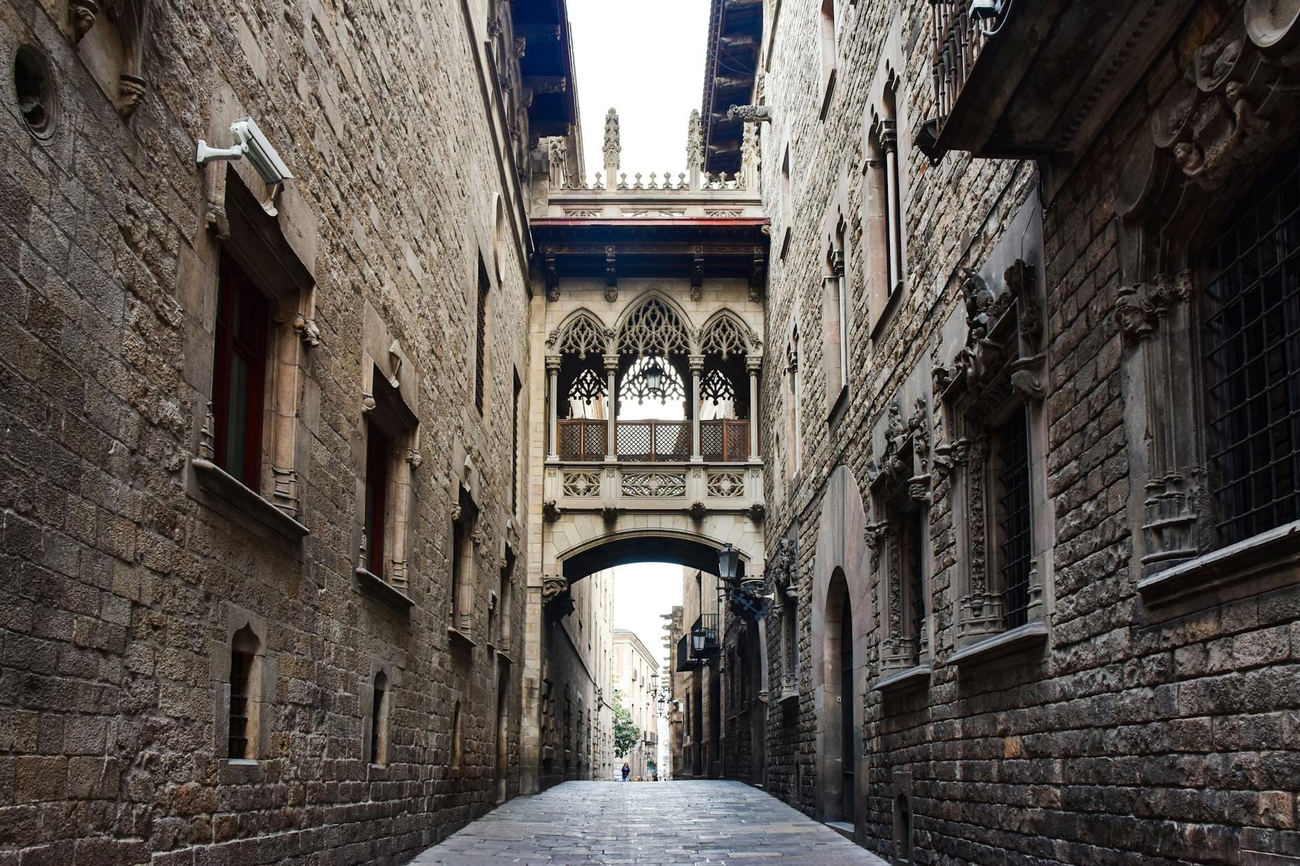 View down Carrer del Bisbe in Barcelona showing the iconic Pont del Bisbe neo-Gothic bridge spanning between two old stone buildings in the Gothic Quarter.